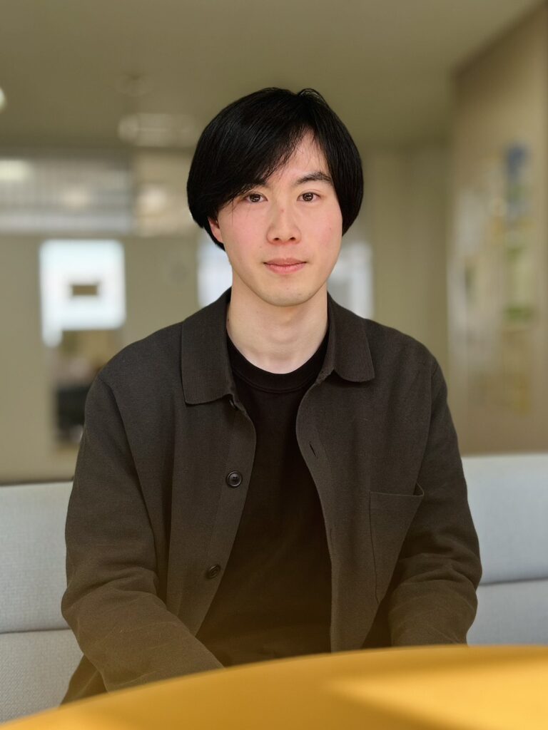 Portrait of a young man with short black hair, wearing a dark shirt and brown cardigan, seated indoors at a table and looking at the camera.