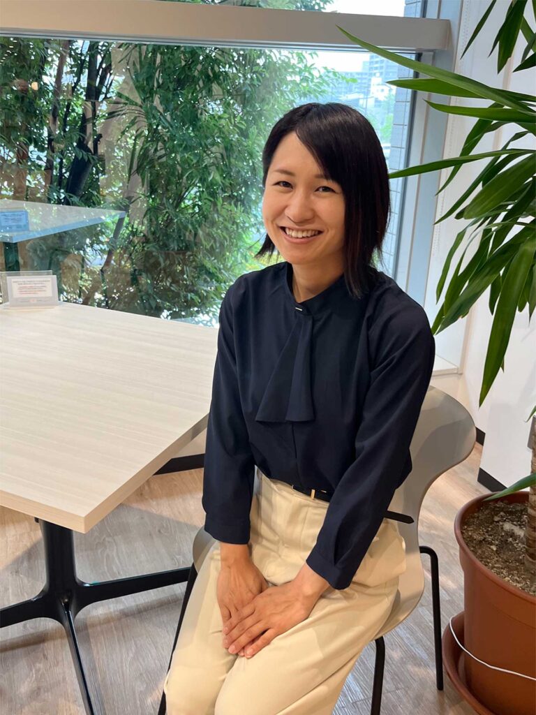 Smiling woman in a black blouse and beige pants sits at a light wood table beside a window with greenery outside.