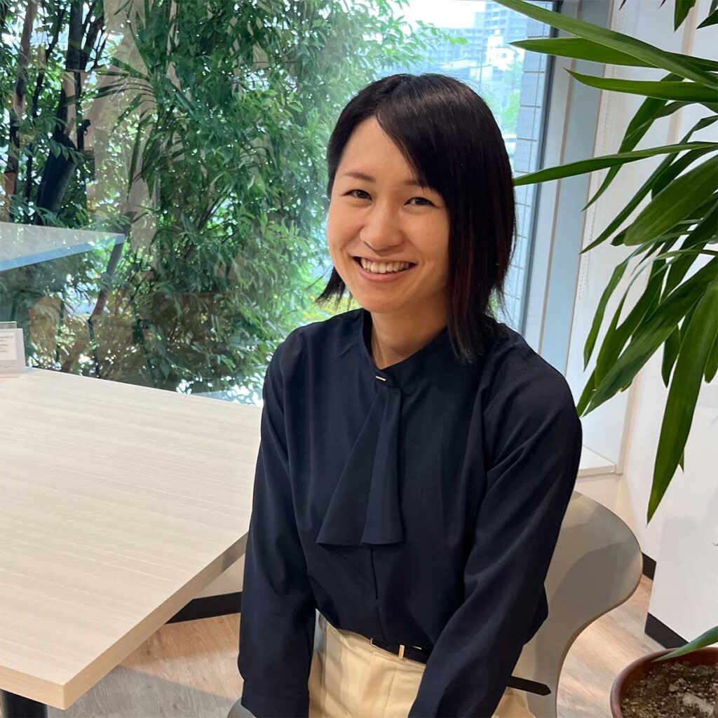 Smiling woman in a navy blouse seated at a light wood table in a bright office with plants visible outside the window.